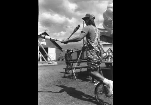 A performer juggles pins while riding a unicycle outside.