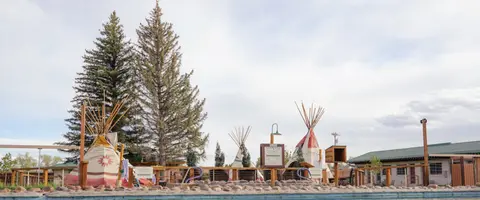 An exterior view of a resort area with tipis, tall trees, and a pool edge in the foreground.