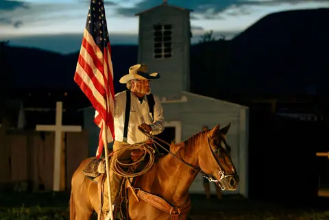 A cowboy holds an American flag while riding a horse in front of a building at dusk.