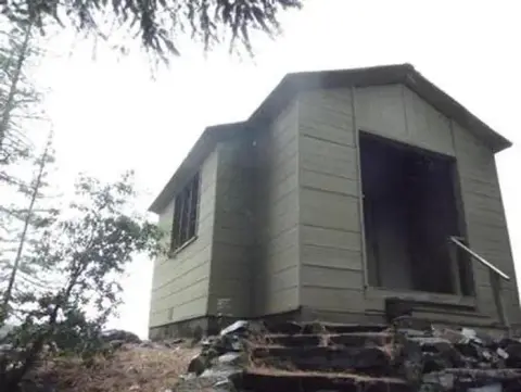 An exterior view of a small building surrounded by trees and rocks.