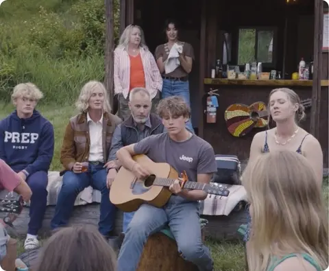 A group of people gathered outdoors listening to someone play guitar.
