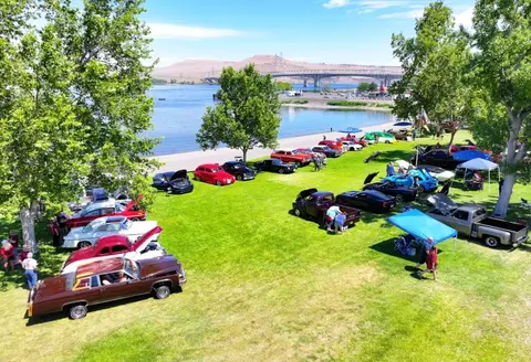 An aerial view shows classic cars parked on grass by a waterfront with a bridge in the background.
