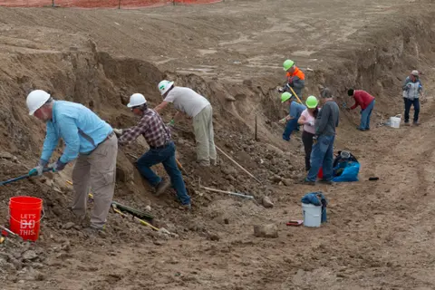 A group of people are digging in a dirt trench with tools.