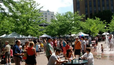 A busy outdoor plaza with people walking around booths and a fountain.