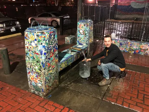 A man filling a glass jug with water from an ornate mosaic fountain at night.