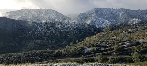 Snow-covered mountains rise behind rolling hills and sparse trees under an overcast sky.