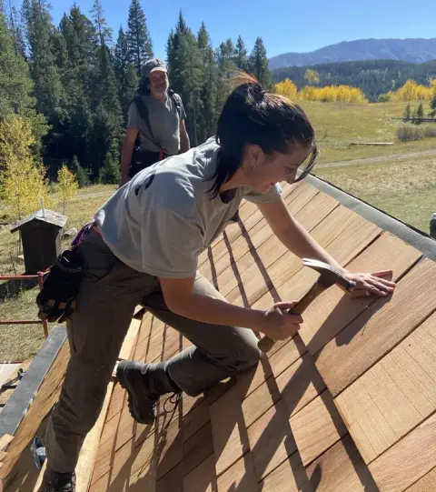 A woman works on installing wood shingles on a roof outdoors.