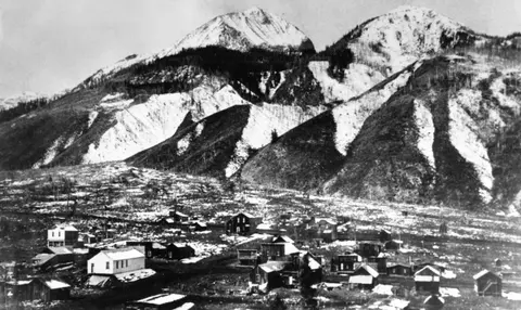 Black and white historic photo showing a small town in front of large snow-capped mountains.