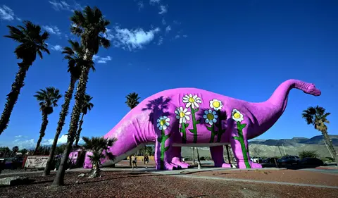 A large pink dinosaur statue decorated with painted flowers stands outdoors under a blue sky.