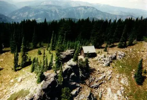 A small cabin sits among rocky terrain and pine trees with mountains in the background.