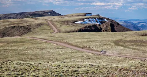A car drives along a dirt road winding through a mountain landscape with patches of snow.