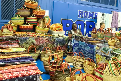A vendor sits behind tables filled with colorful fabrics and woven baskets at an indoor market.
