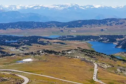 An aerial view of a winding mountain road with lakes and distant snow-capped peaks.
