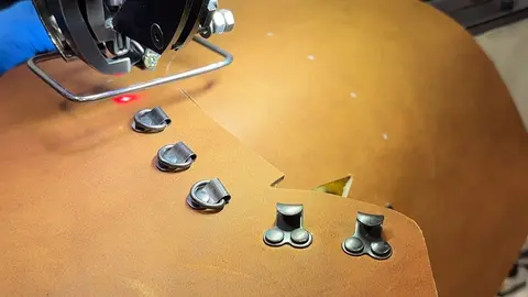A machine cuts brown leather near metal hardware attached to the material.