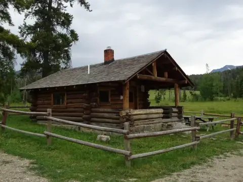 A log cabin with a fenced yard stands in a grassy area with trees and mountains.