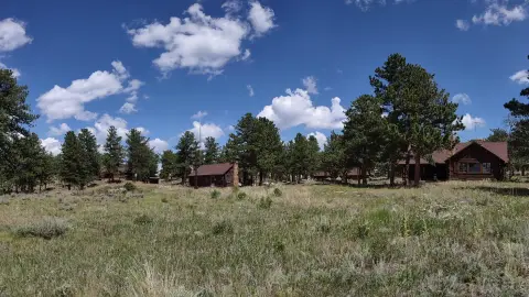 A panoramic view of wooden cabins set amidst grassy fields and pine trees under a blue sky with white clouds.
