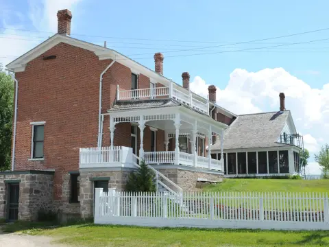 A historic brick farmhouse with a white porch and fence under a blue sky.