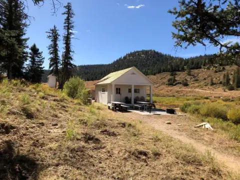 A small white cabin sits in a mountain clearing with picnic tables outside.