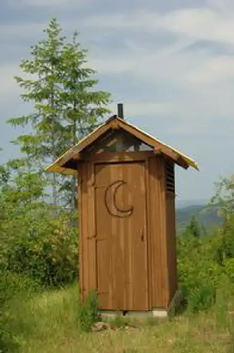 An outdoor wooden outhouse stands in a grassy area with trees.