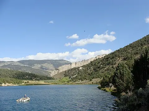 A person rows a small boat on a lake surrounded by mountains and trees.