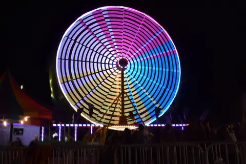 A brightly lit Ferris wheel spins at night with people gathered in front of it.