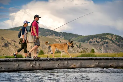 Two people and a dog walk across a wooden footbridge holding fishing rods.