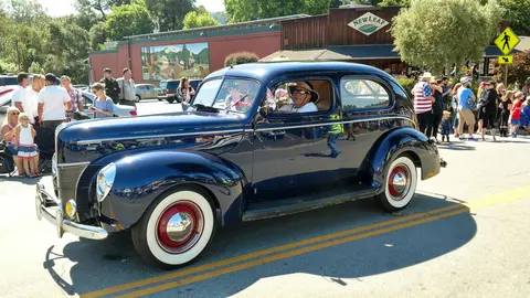 A dark blue vintage car drives down a street lined with people and buildings.