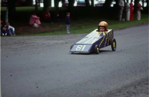 A person races a soapbox car down a paved track.