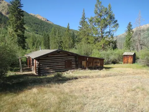 A log cabin in a grassy field with mountains and trees in the background.