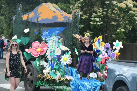 A child in costume rides on a parade float decorated with giant flowers and a mountain backdrop.