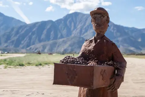 A metal sculpture of a farmworker carrying a crate of grapes outdoors.