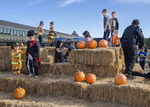 Children in costumes play on a large stack of hay bales with pumpkins around a train.