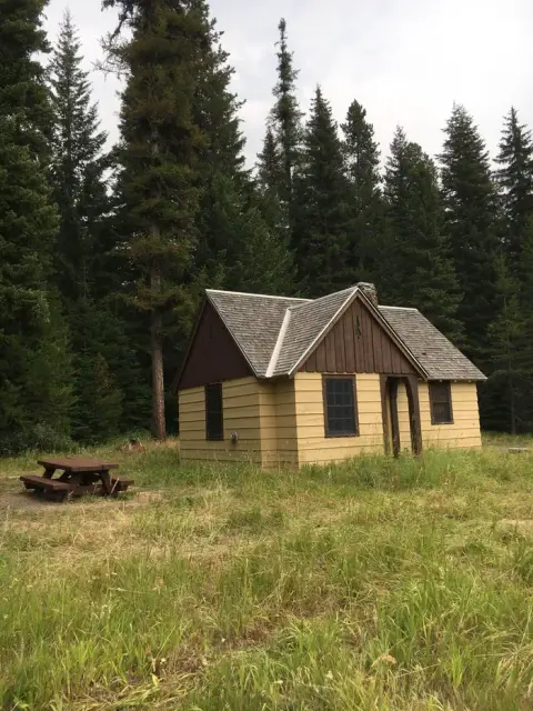 A small cabin stands in a clearing surrounded by tall evergreen trees.