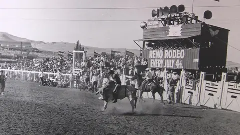 Black and white historic photo of a rodeo with a bull rider and spectators.