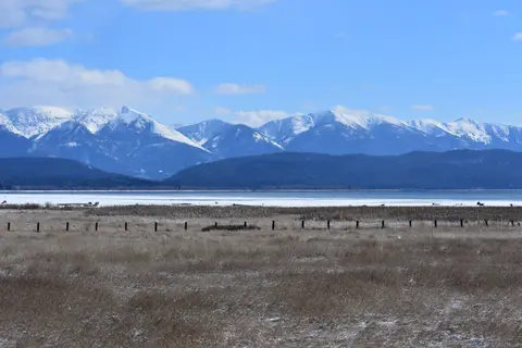 A scenic view of snow-covered mountains, a lake, and a field under a blue sky.
