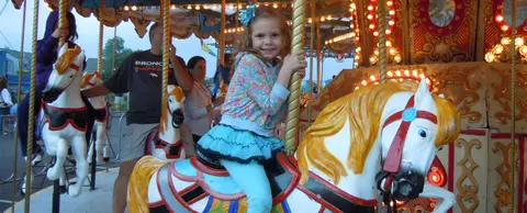 A young girl rides a carousel horse while smiling at the camera.
