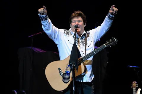 A musician performs on stage holding a guitar and pointing up with both hands.