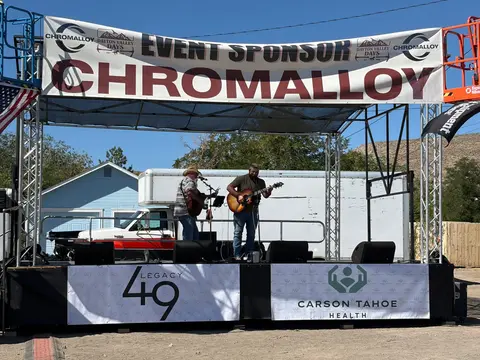 Two musicians perform on an outdoor stage with sponsor banners.