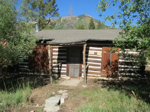 An old log cabin sits in a clearing with a mountain and trees in the background.