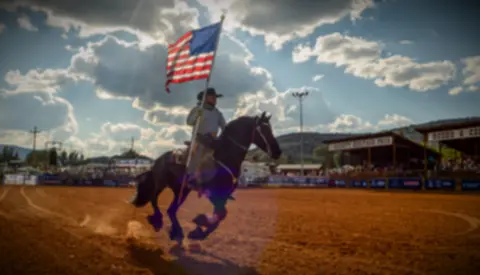 A rider carrying an American flag rides a horse across a dirt arena under a cloudy sky.
