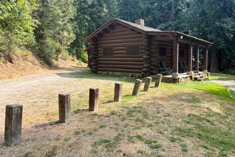 A log cabin sits in a clearing surrounded by trees, with a line of wooden posts in the foreground.