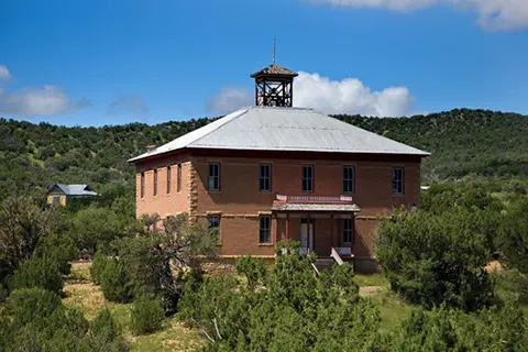 A historic two-story brick building with a cupola stands among green trees and hills.
