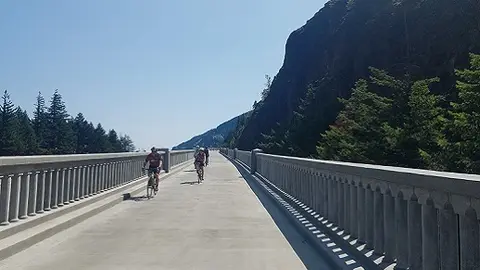 Cyclists ride across a wide concrete bridge flanked by stone railings.