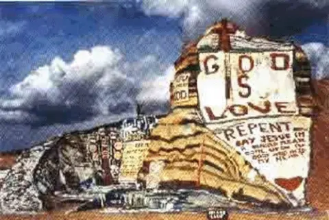 A photograph of a painted rock structure with religious messages in a desert setting.