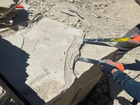 A person using chisels to split a large flat stone slab outdoors.