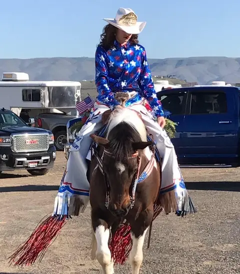 A person in patriotic western attire rides a horse outdoors.