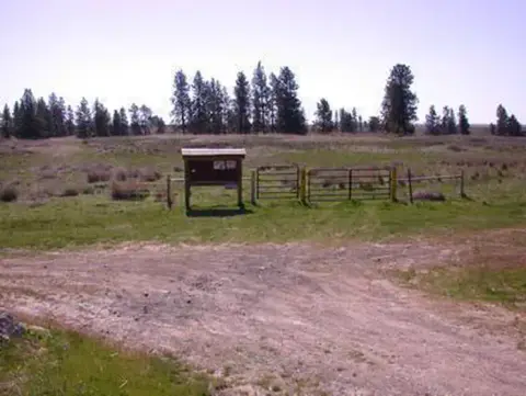 A dirt road leading to an open field with a sign board and fence.