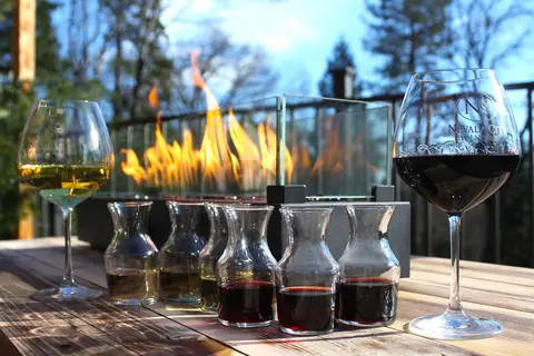 Glasses and carafes of wine sit on a wooden table outdoors in front of a fire pit.