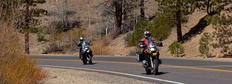 Two motorcyclists ride along a winding road through a mountainous landscape.