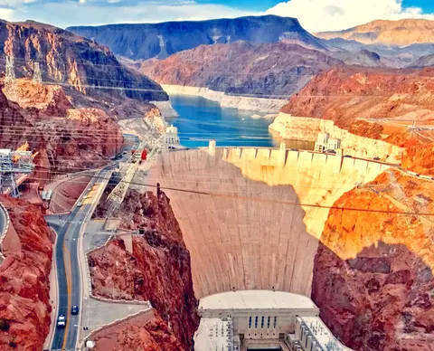 An aerial view of a large concrete dam set between rocky canyons with a blue reservoir behind it.